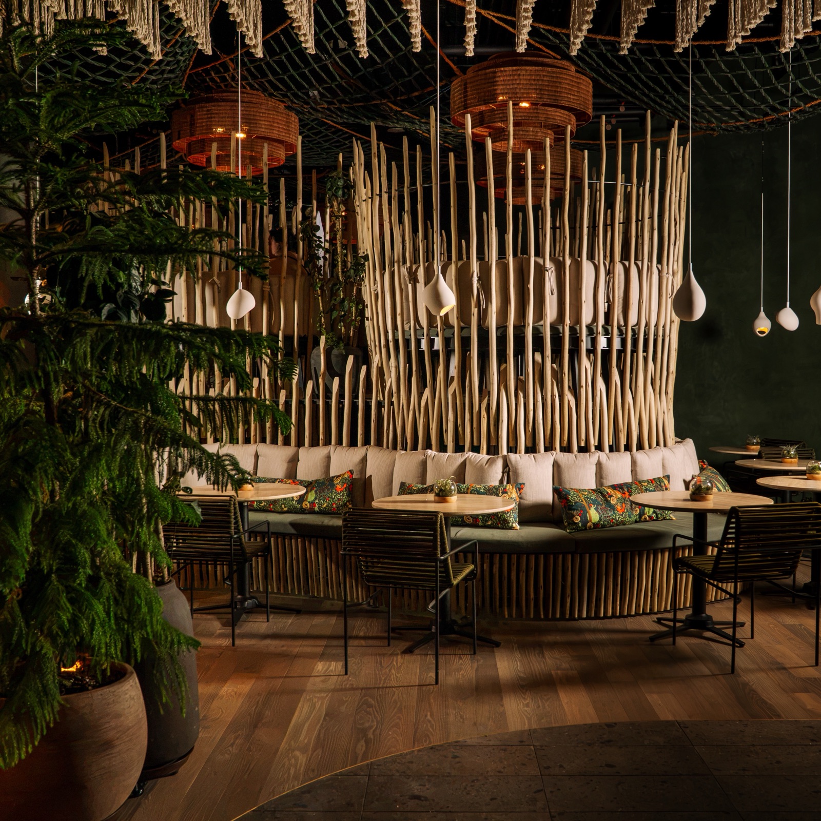 A curved green banquette at Forbidden Fruit, set beneath a sculptural canopy of hanging carved paddles and woven baskets, with large potted ferns on the timber floor.