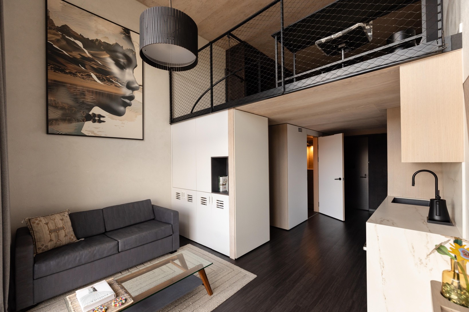 The living level of a Sky Loft, with a grey sofa and low timber table under a double-height wall, a black mesh mezzanine rail overhead and a small kitchen with a stone counter to the right.