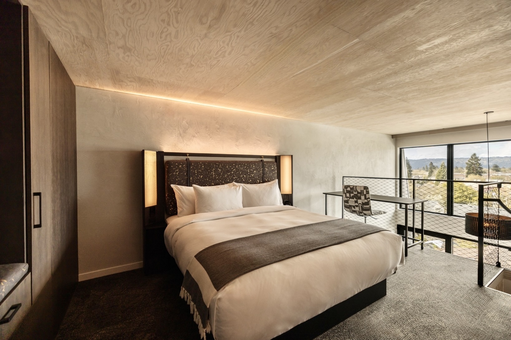 A Sky Loft sleeping platform under a raw plywood ceiling and bare concrete wall, the bed lit by two slim wall lamps with a corner window opening onto treetops and hills.