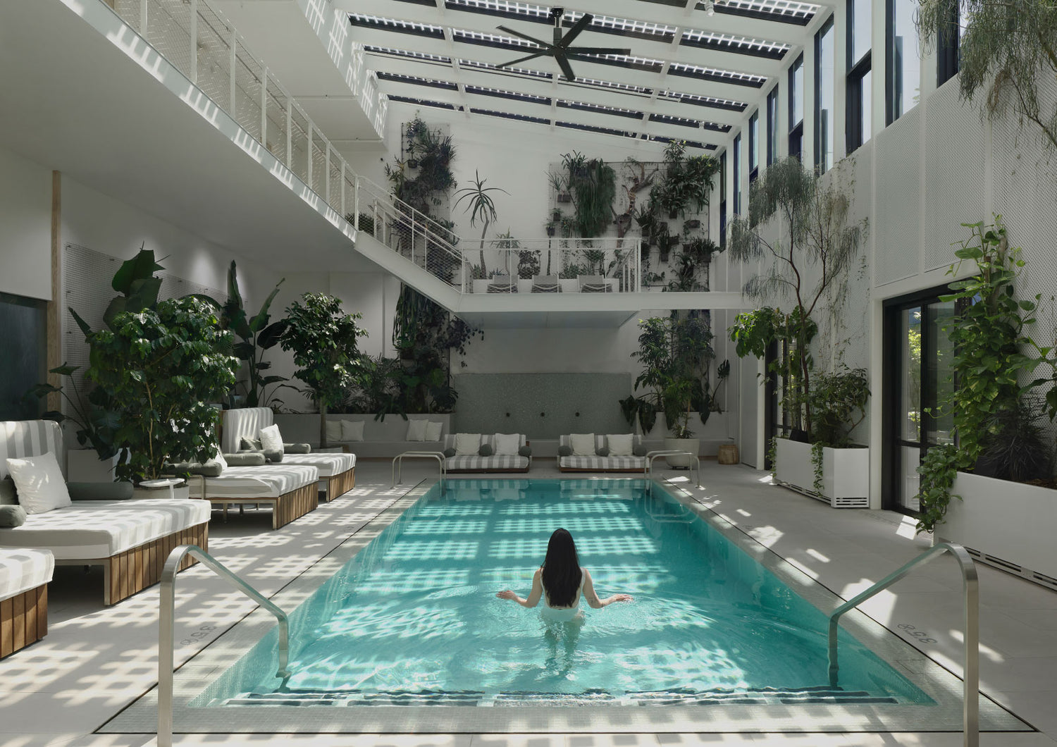 The Conservatory pool at Cascada, a long turquoise bath set in a tall white atrium under a slatted glass roof, ringed by loungers and banked planters of ferns and palms, with one swimmer standing at the far end.